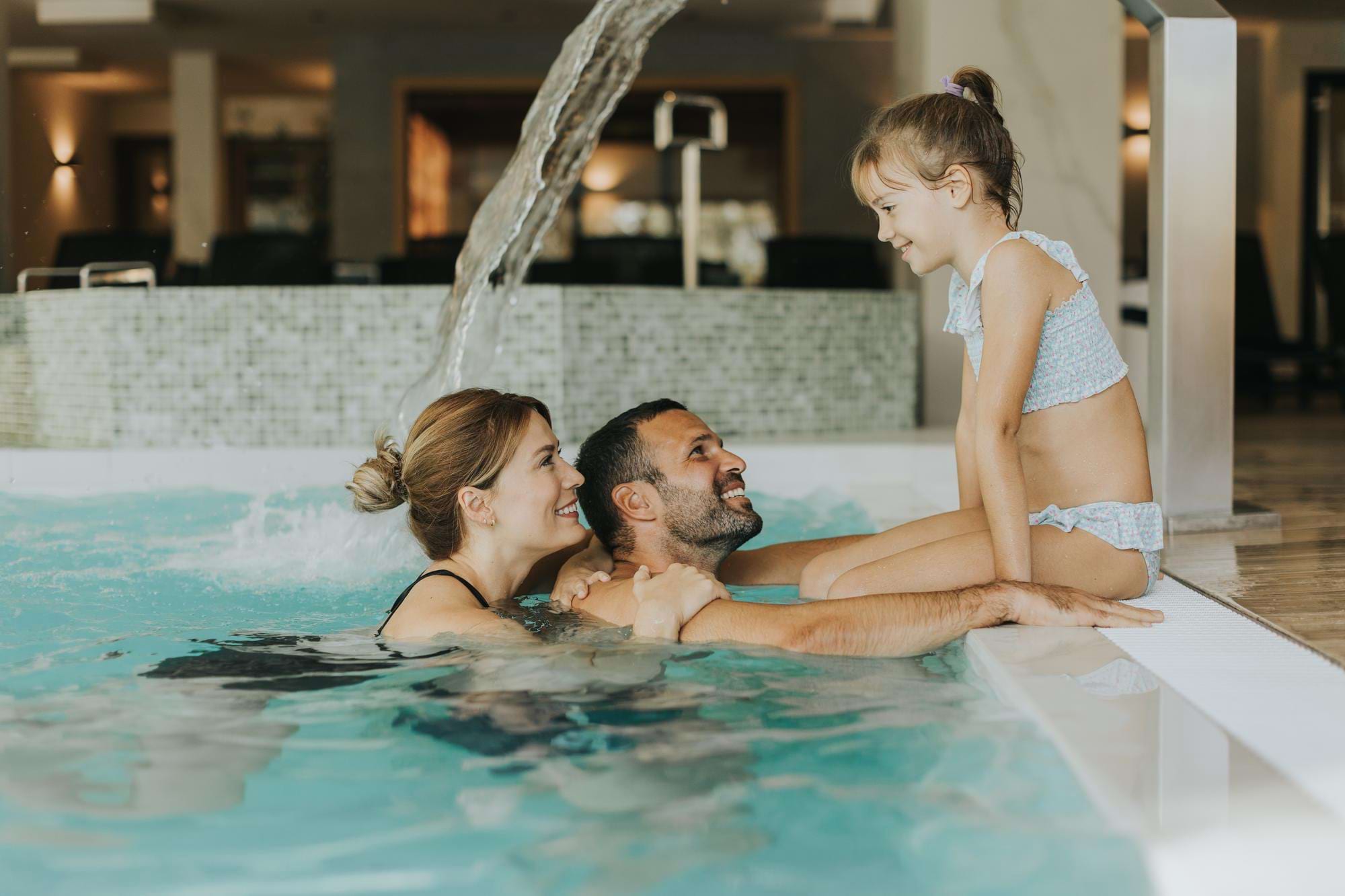 Dziewczynka w bikini na brzegu basenu, rodzice w wodzie uśmiechają się