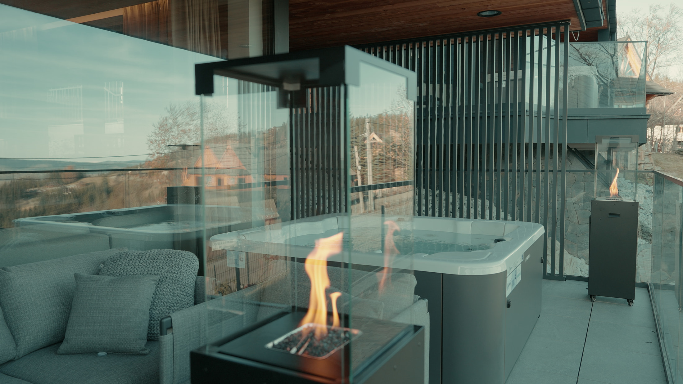 Taras z jacuzzi, palenisko płonie, szklane balustrady i sofa